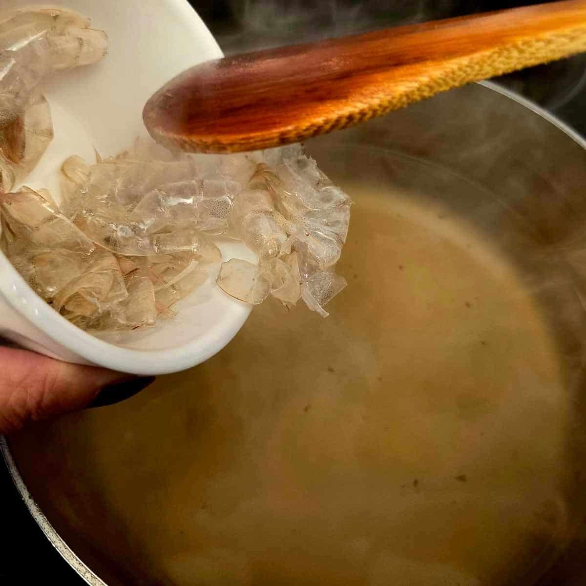 Hands adding shrimp shells to a pot of simmering broth on a stove