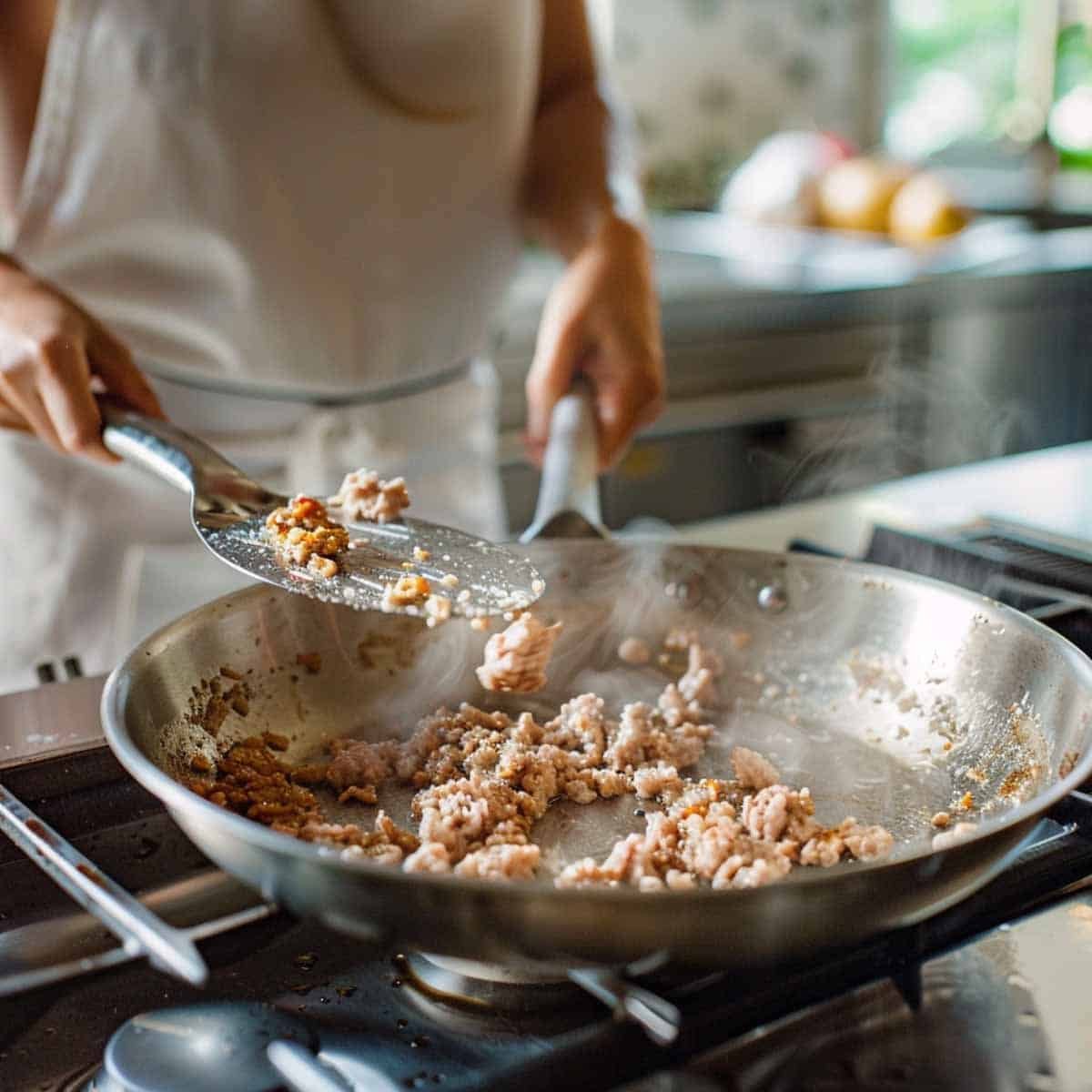 Minced pork sizzling in a stainless steel pan, being cooked for Larb, also known as Laab. The pork is being stirred with a stainless steel spoon, with visible steam rising, and seasonings like lime juice and herbs nearby on the countertop.