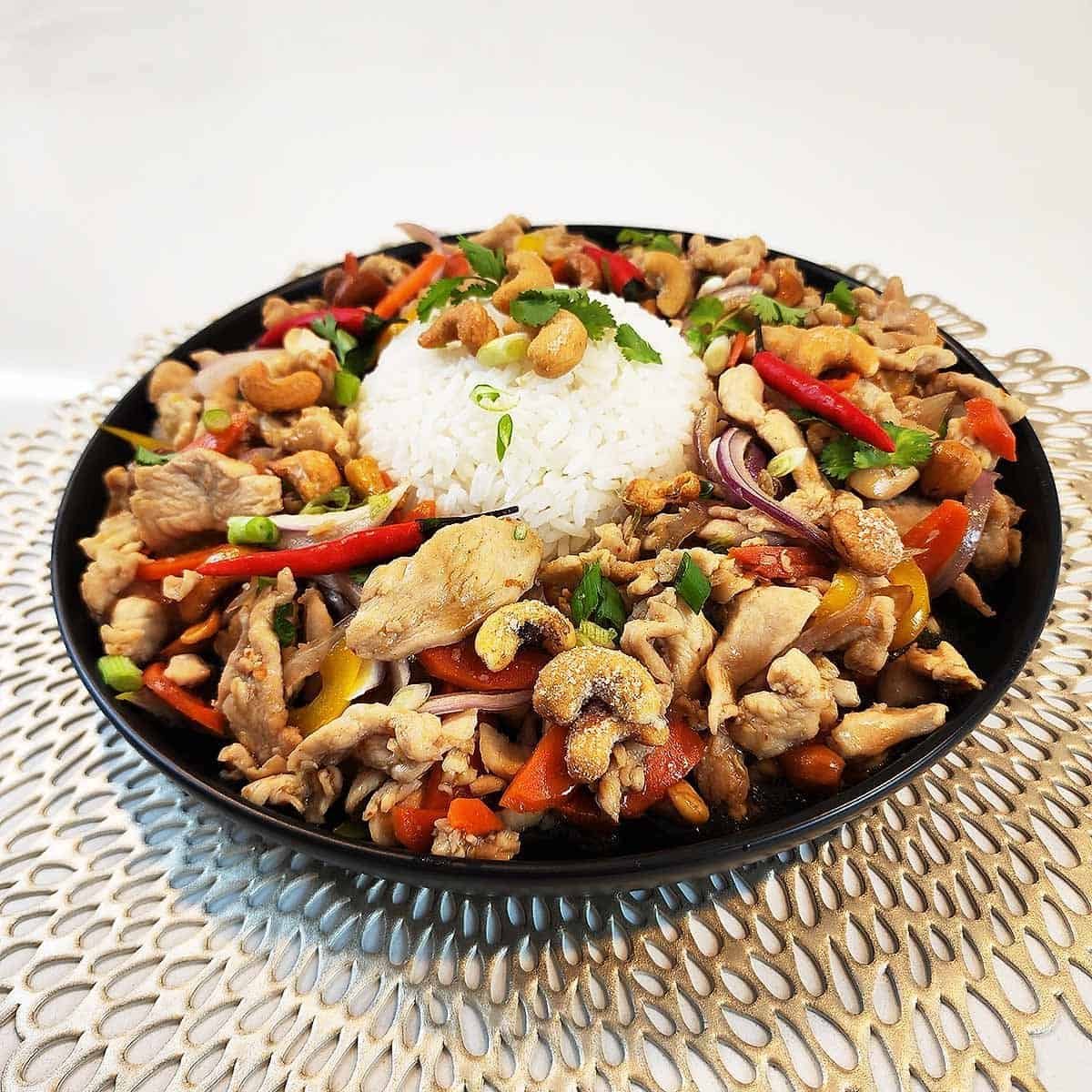 Stir-fried chicken pieces with roasted cashews, colorful bell peppers, and green onions served over steamed jasmine rice in a white bowl