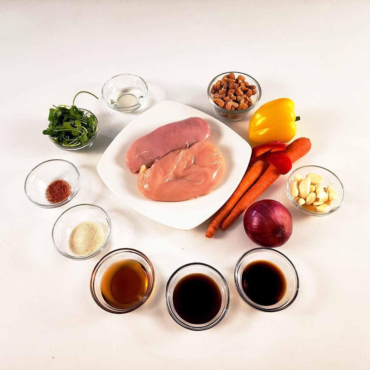 Fresh ingredients for Thai Cashew Chicken spread on white countertop: chicken, cashews, vegetables, and Asian sauces