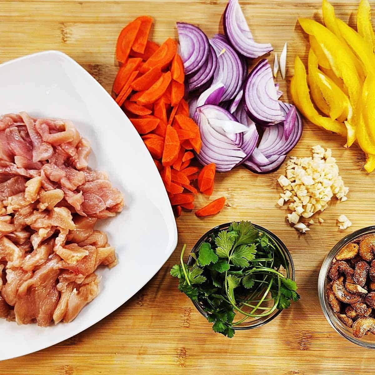 Raw ingredients for Thai Cashew Chicken arranged on bamboo cutting board: sliced chicken breast, cashews, bell peppers, green onions, garlic, and Thai chilies