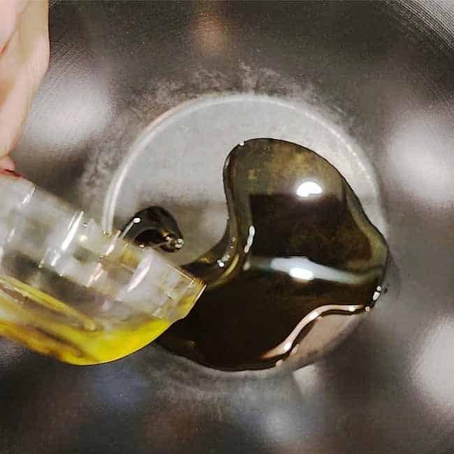 Hand pouring cooking oil from bottle into heated wok with steam rising