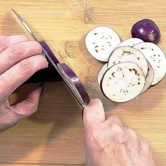 Purple eggplant being sliced on a bamboo cutting board with a chef's knife