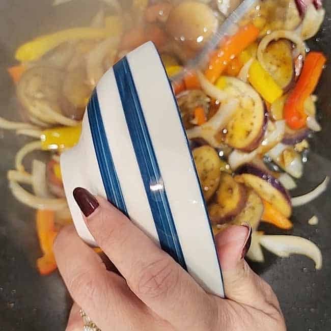 Sauce being poured from small bowl over sizzling vegetables in a hot wok