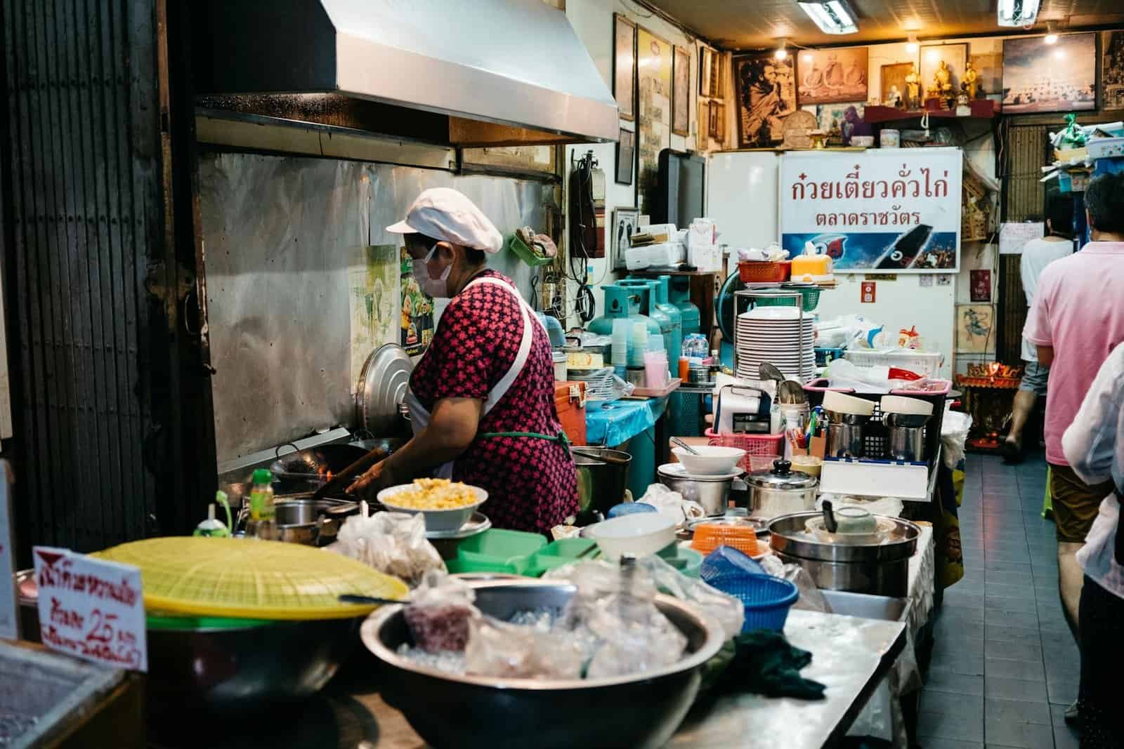 Street food vendor in a bustling Bangkok market kitchen preparing dishes with fresh ingredients.